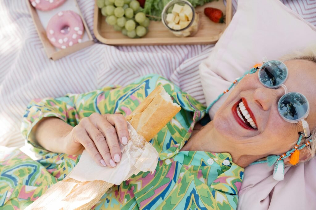 Cheerful elderly woman in colorful attire enjoys a picnic with baguette, donuts, and fresh fruits outdoors.